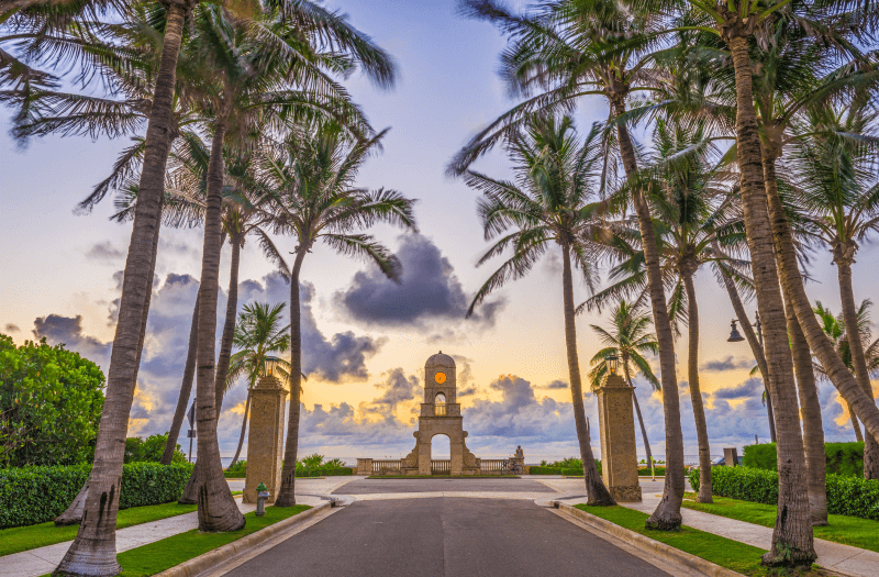 View of a road surrounded by palm trees with sidewalks on the sides, with a clock and sunset sky in the background