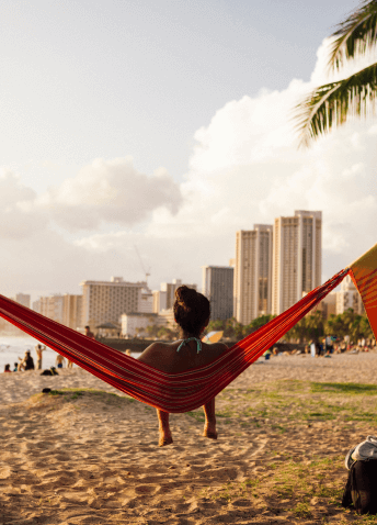 View from behind of a person sitting in a hammock on the beach, with buildings and sunset sky in the background