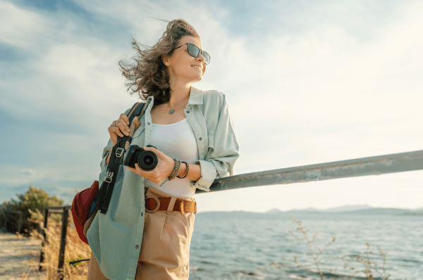 Person leaning on a railing, holding a camera with the sea and vegetation in the background