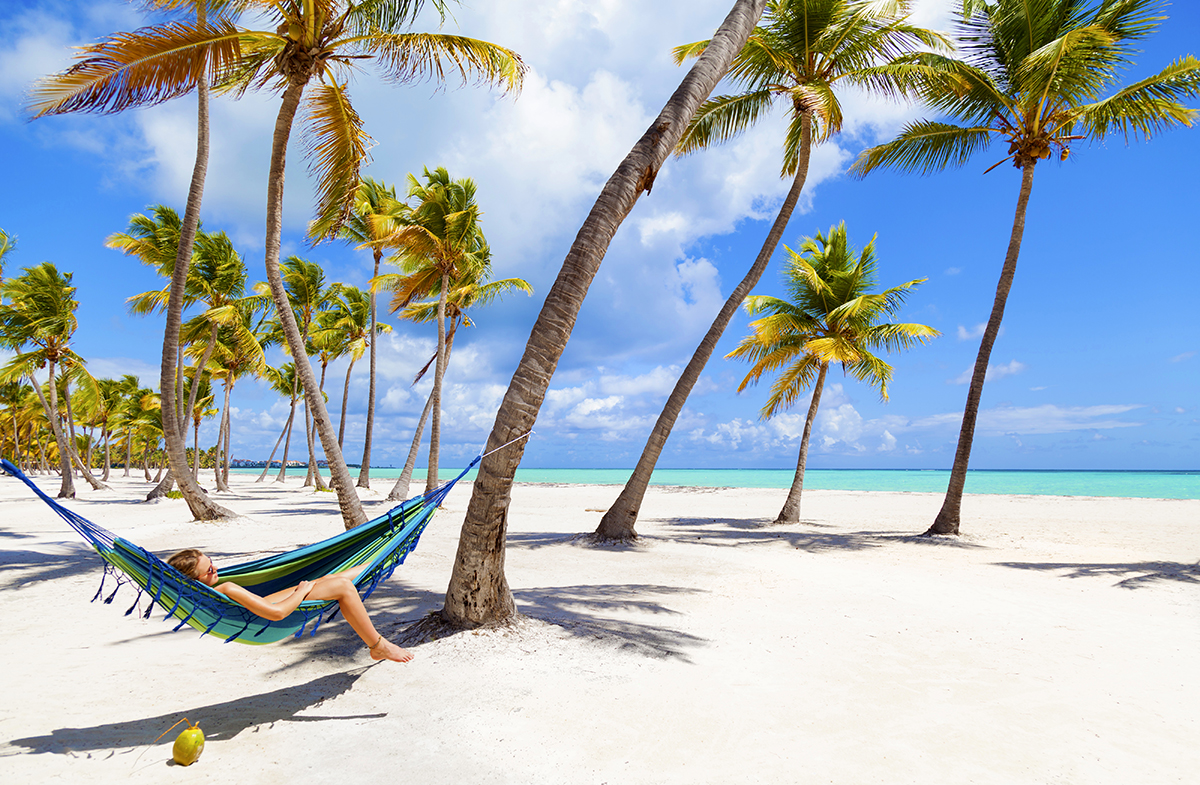 A young woman lounging on a hammock on a sandy beach with palm trees.