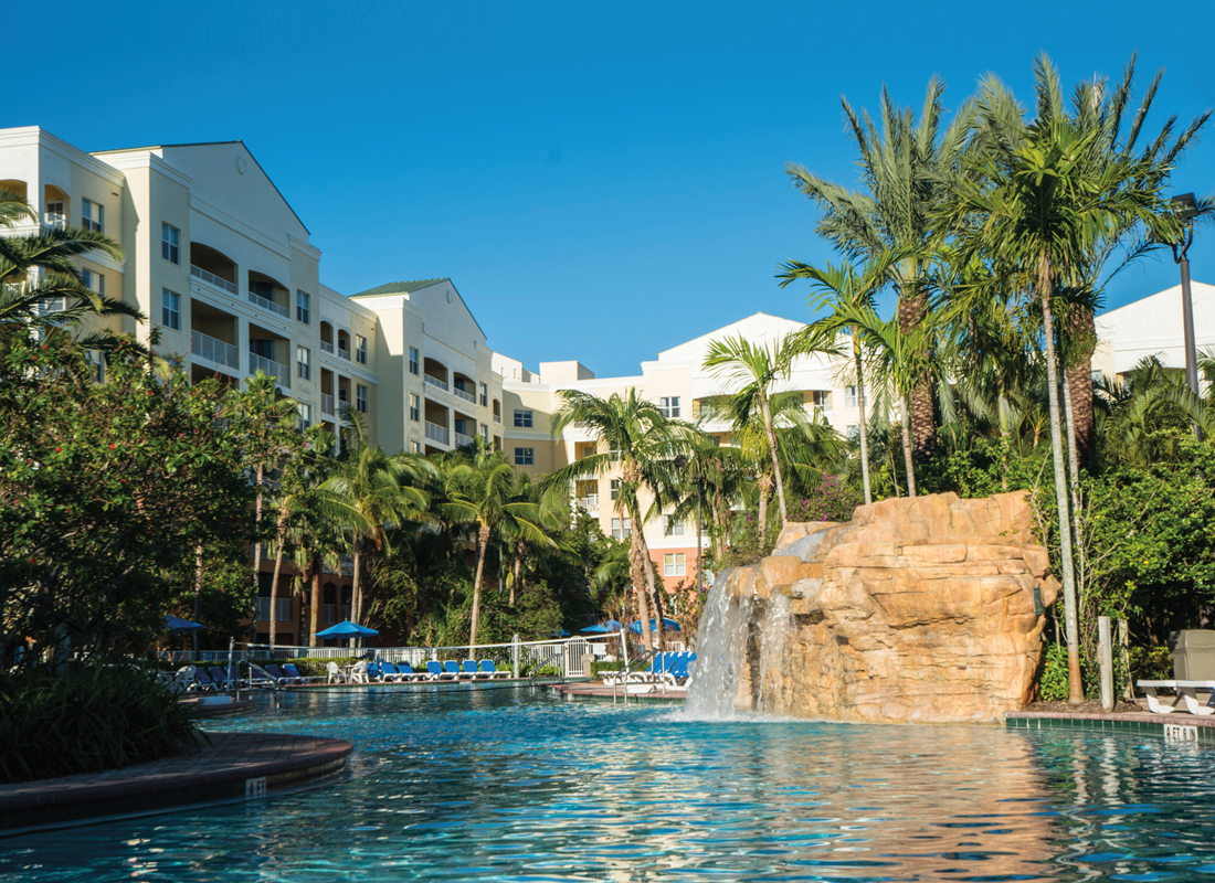 View of a swimming pool with a waterfall, surrounded by palm trees, buildings and with a clear sky in the background