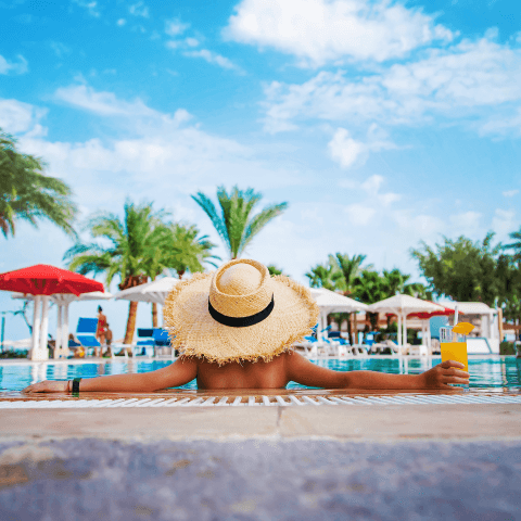 Person with hat in swimming pool holding a drink, with palm trees, umbrellas and sky with clouds in the background