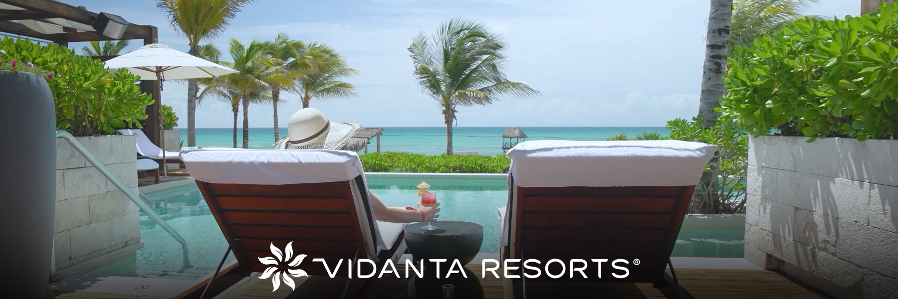 View from behind of a person lying on a lounge chair and holding a drink in front of a swimming pool, with palm trees and the sea in the background