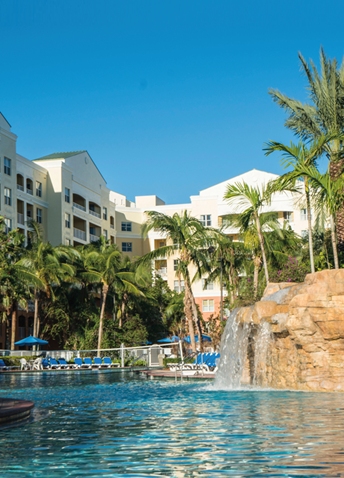 View of a pool with a waterfall and surrounded by buildings and palm trees, with a clear sky in the background