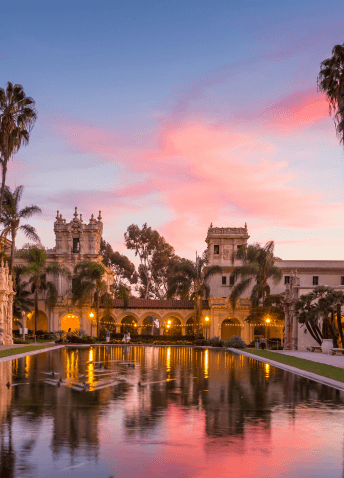 View of a pond surrounded by palm trees, with old building and sunset sky in the background